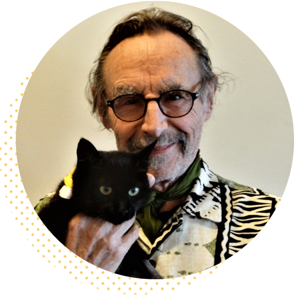 Elderly man wearing a patterned shirt and glasses smiles while holding a black cat with green eyes, set against a neutral background.