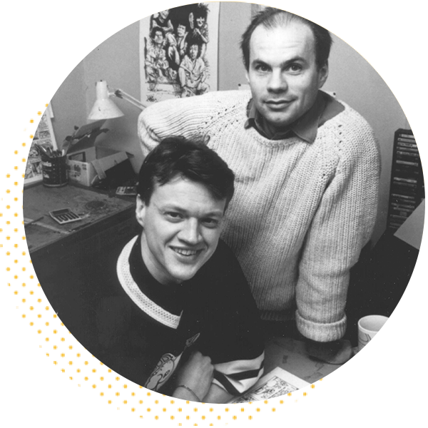 Black and white photo of two men in an office setting, one sitting at a desk and the other standing behind him. The desk is cluttered with art supplies and papers, showcasing a creative workspace atmosphere.