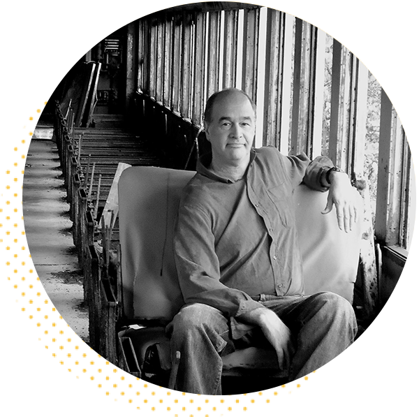 Portrait of a man seated in an industrial setting, wearing a button-up shirt and jeans, with a relaxed expression. The background features a long corridor with large windows, suggesting an abandoned warehouse or factory. The image is in black and white, highlighting the contrast between the subject and the surroundings.