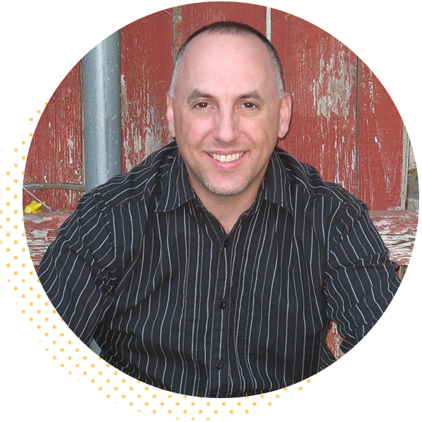 Portrait of a smiling man with short hair wearing a black striped shirt, seated against a rustic red wooden background, conveying a friendly and approachable demeanor.