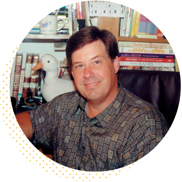 Portrait of a smiling man in a patterned shirt sitting at a desk with a white duck decoration and a background filled with books and office supplies.