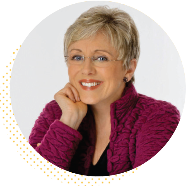 Smiling woman with short blonde hair and glasses, wearing a purple sweater, resting her chin on her hand against a white background.