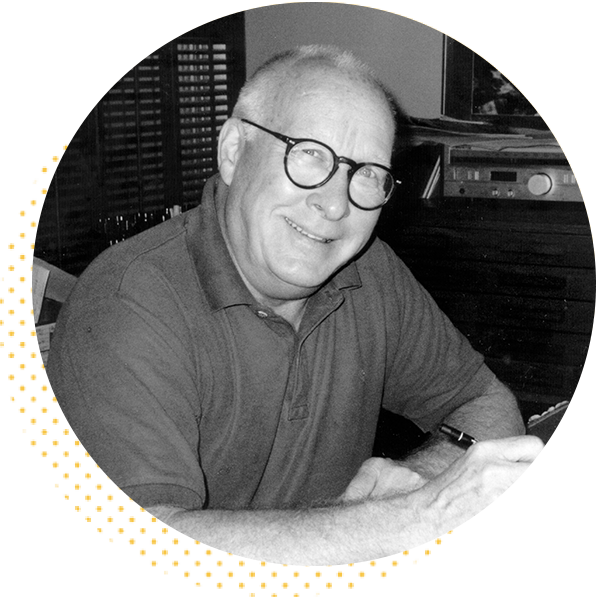 Smiling older man with glasses sitting at a desk, engaged in writing or working on a project, surrounded by shelves and a vintage radio.