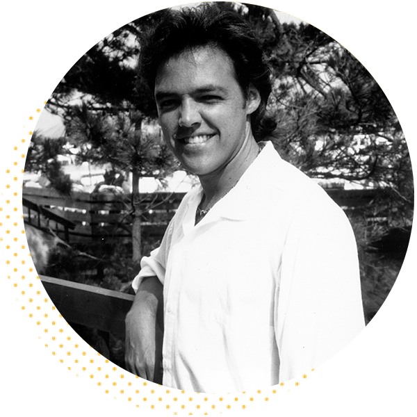 Black and white portrait of a smiling man in a white shirt, standing outdoors with trees in the background, showcasing a friendly and approachable demeanor.