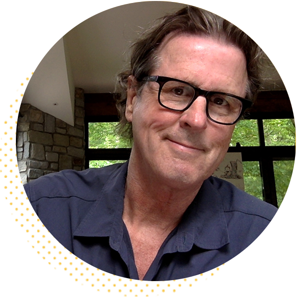 Smiling man with glasses in a casual shirt, seated indoors with a stone wall backdrop and green trees visible through large windows.
