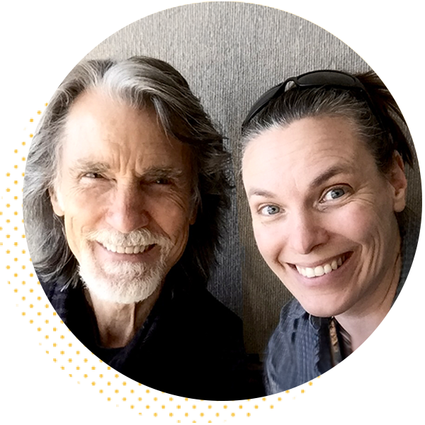 Smiling man with gray hair and beard alongside a woman with dark hair in a casual setting, both posing for a selfie. The background features a neutral-colored wall, highlighting their joyful expressions.