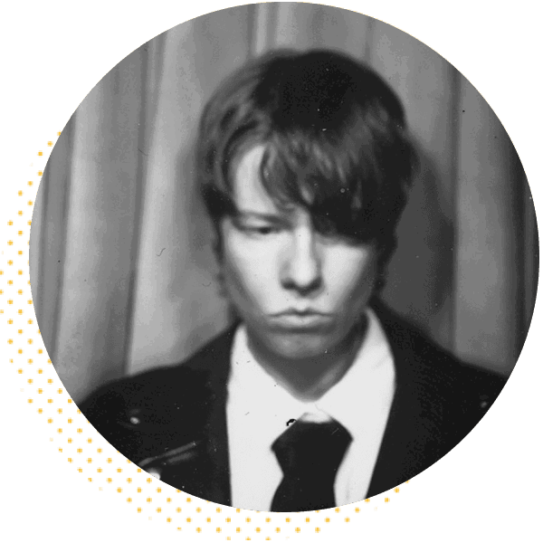 Black and white photo booth image of a young man with medium-length hair, wearing a suit and tie, looking downward with a solemn expression against a curtain backdrop.