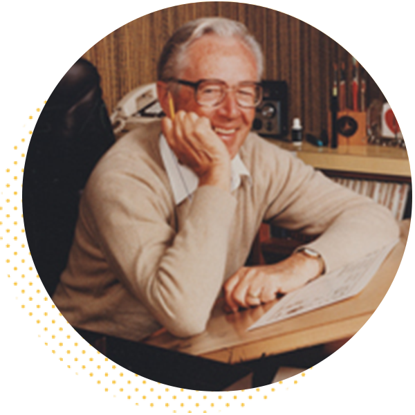 Smiling elderly man in a beige sweater resting his chin on his hand, seated at a desk with drawing materials and vintage items in the background. The image captures a warm, creative atmosphere.