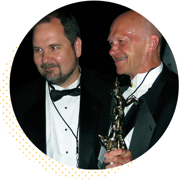 Two men in tuxedos smiling at an awards event, one holding a trophy. The scene captures a moment of celebration and achievement in a formal setting.