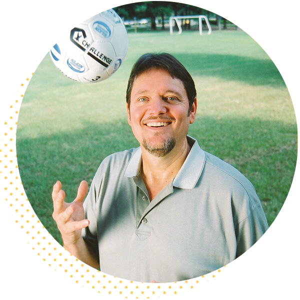 Smiling man in a gray polo shirt juggling a soccer ball in a park setting, with soccer goals visible in the background.