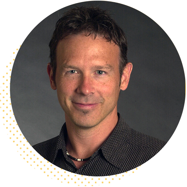 Portrait of a smiling man with short hair, wearing a patterned shirt and necklace, set against a dark background. The image is circular and highlights his approachable demeanor.