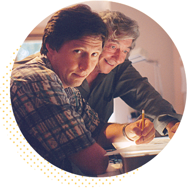 Two men engaged in a creative work session, one looking directly at the camera and the other writing notes. They are surrounded by a workspace filled with design materials, reflecting collaboration and artistic focus.