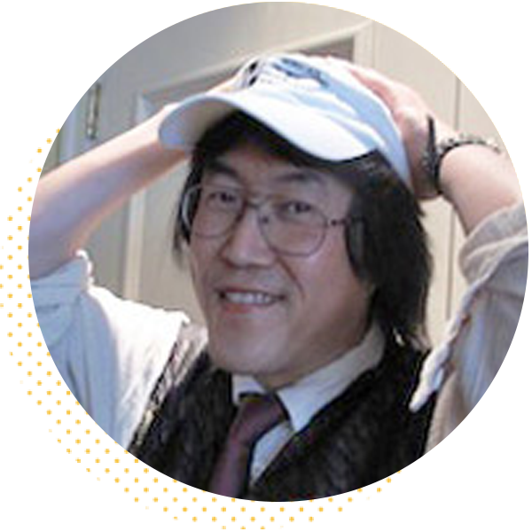 Smiling man with glasses and long black hair, wearing a white cap and a vest, poses with his hands on his head against a light background.
