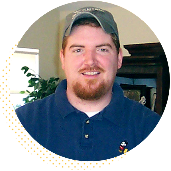 Smiling man with a beard wearing a blue polo shirt and baseball cap, standing indoors with plants and furniture in the background.