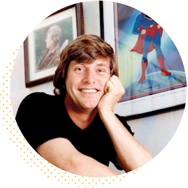 Smiling man resting his chin on his hand, seated in front of framed artwork, including a Superman poster, exuding a cheerful demeanor.