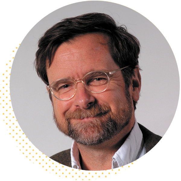 Smiling man with glasses and a beard, wearing a white shirt and a brown jacket, against a neutral background.