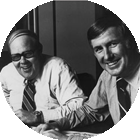 Two men smiling and engaged in conversation at a table, wearing formal attire with ties, in a professional office setting. The image captures a moment of collaboration or discussion.