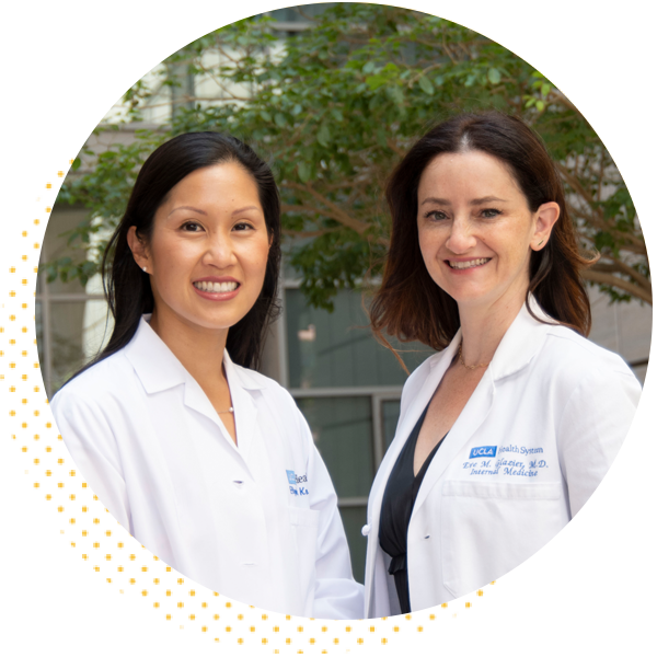Two female healthcare professionals in white lab coats stand together outdoors, smiling against a backdrop of greenery, representing teamwork and medical expertise.