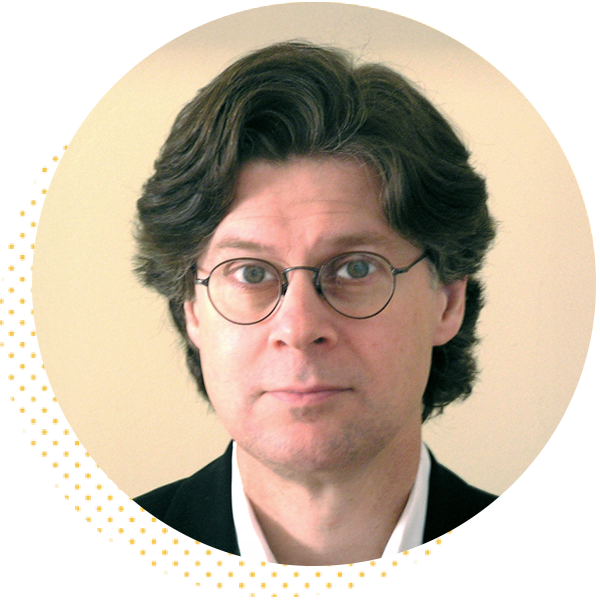 Portrait of a man with medium-length wavy hair and round glasses, wearing a black blazer and white shirt, set against a plain background. Ideal for professional or academic contexts.