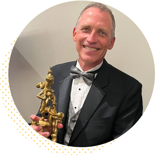 Smiling man in a tuxedo holding a gold trophy featuring a stacked human figure, set against a neutral background.
