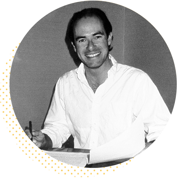 Smiling man in a white shirt sitting at a desk with papers, holding a pen, in a black and white photograph. Ideal for showcasing creativity and professional engagement.