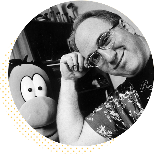 Black and white image of a smiling man with glasses, wearing a colorful shirt, posing playfully next to a cartoon character. The background features shelves with various items, adding a creative atmosphere.