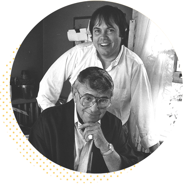 Two men pose together in a cozy indoor setting, with one man seated in front, wearing glasses and a thoughtful expression, while the other stands behind him smiling. The image captures a moment of camaraderie and creativity, highlighting their connection in a professional or artistic environment.