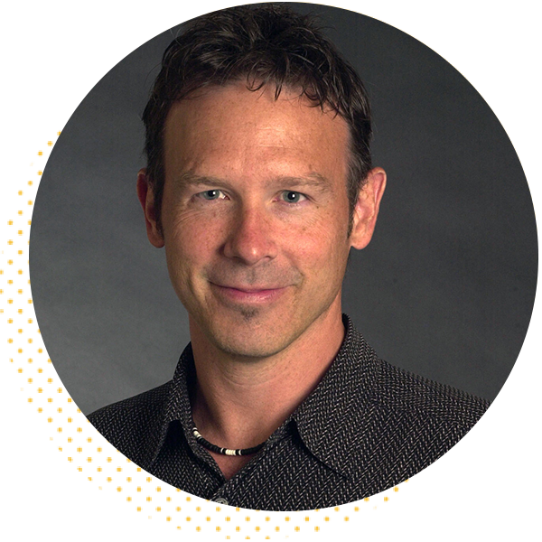 Professional headshot of a man with short, dark hair, wearing a patterned shirt and a necklace, set against a neutral background.