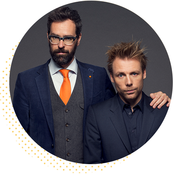 Two men in formal attire posing for a portrait, one with glasses and a beard wearing a blue suit and orange tie, the other with tousled hair in a dark suit. The background is a circular design with a subtle pattern.