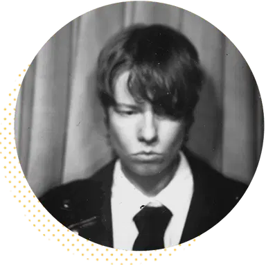 Black and white photo booth portrait of a young person with tousled hair, wearing a suit and tie, looking serious against a curtain backdrop.