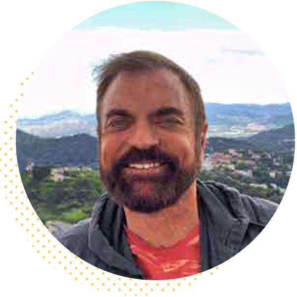 Smiling man with a beard wearing a gray jacket, standing outdoors with a scenic mountainous background. The image captures a sunny day with clear skies and greenery, reflecting a joyful and relaxed atmosphere.