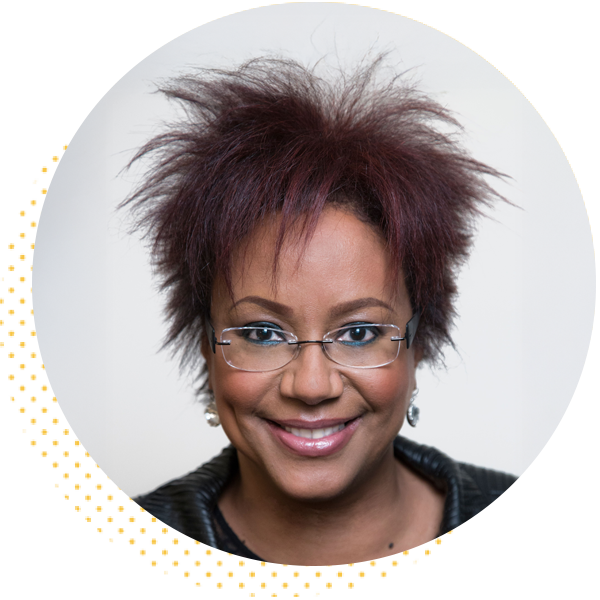 Professional headshot of a smiling woman with short, spiky red hair and glasses, wearing a black jacket. The image features a circular crop with a soft background, highlighting her friendly expression and approachable demeanor.