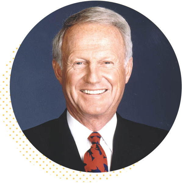 Smiling middle-aged man with short gray hair, wearing a black suit and a red tie with blue patterns, against a dark blue background.