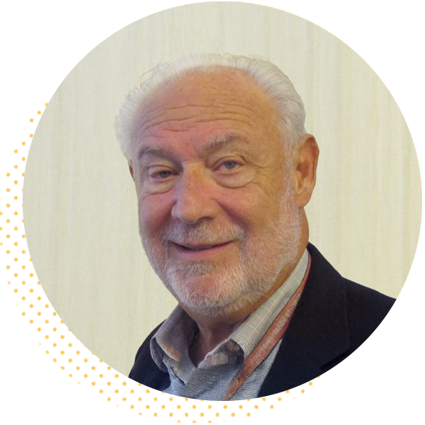 Smiling older man with a beard wearing a blazer and patterned shirt, set against a light background, showcasing a friendly demeanor.