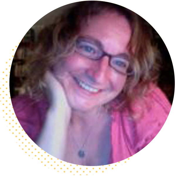 Smiling woman with curly hair and glasses resting her chin on her hand, wearing a pink shirt, against a blurred background.