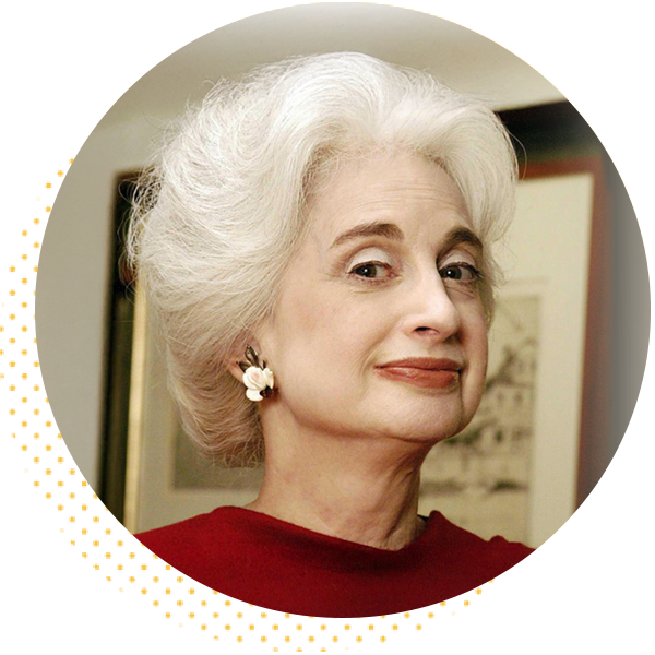 Portrait of an older woman with elegant white hair styled in soft waves, wearing a red top and pearl earrings, smiling confidently against a blurred background of framed artwork.