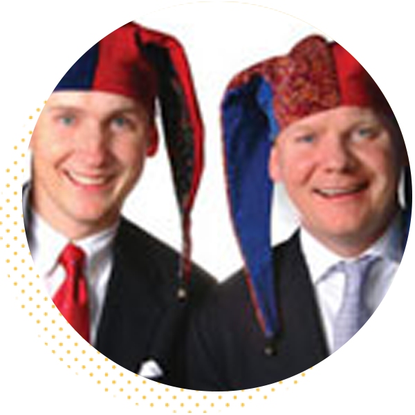 Two men smiling while wearing colorful jester hats, dressed in formal attire, showcasing a playful and festive vibe.