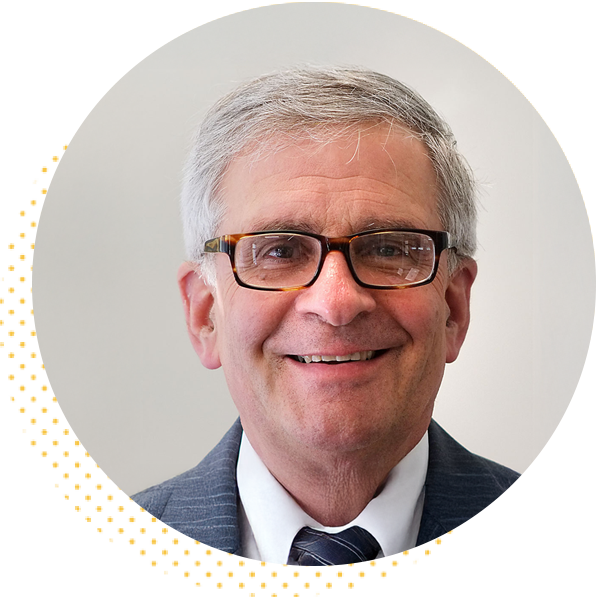 Smiling professional man with glasses and gray hair, wearing a suit, against a light background.