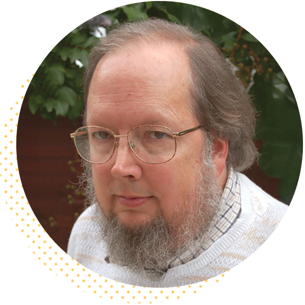 Portrait of a man with glasses and a beard, wearing a light sweater, set against a backdrop of green foliage.