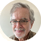 Portrait of an older man with gray hair and glasses, smiling warmly against a neutral background, highlighting a friendly and approachable demeanor.