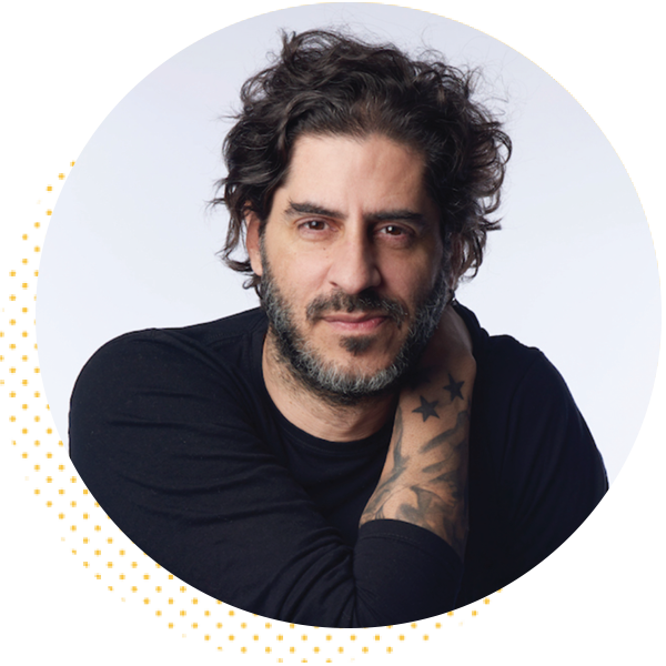 Portrait of a man with curly hair and a beard, wearing a black long-sleeve shirt, showcasing a tattoo on his arm. The background is softly lit, enhancing the subject's features.