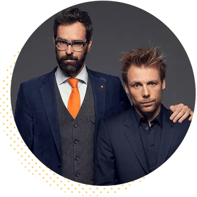 Two well-dressed men stand closely together, one in a suit with glasses and an orange tie, and the other in a dark shirt. They exude a confident and professional demeanor against a dark background.