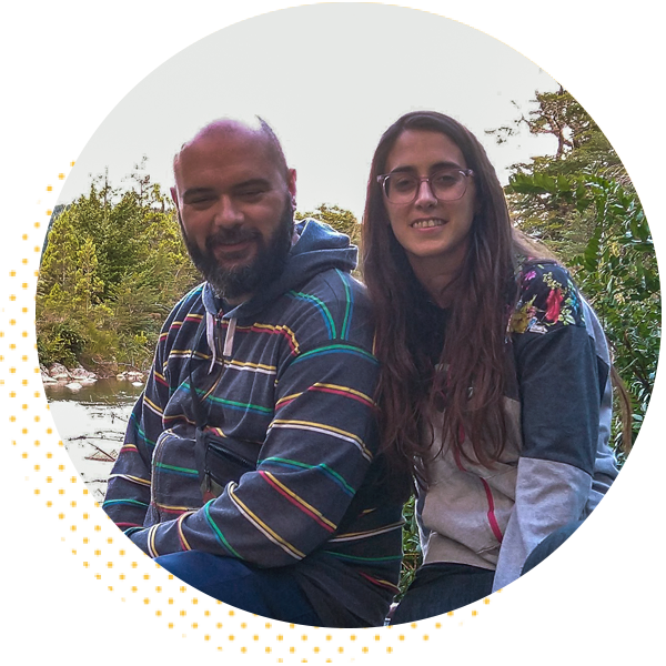 A man and a woman sitting together by a river, surrounded by greenery. The man has a beard and is wearing a striped hoodie, while the woman is wearing a gray jacket and glasses.